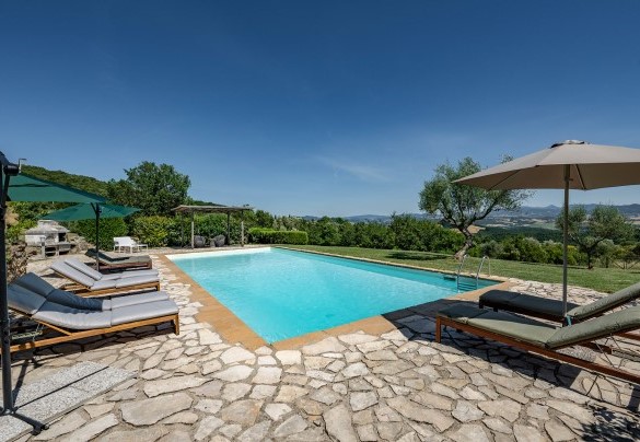 Su una superficie in pietra sono disposti lettini, un divano con poltrone ombreggiati da un gazebo ed un ombrellone – Cinque Cipressi