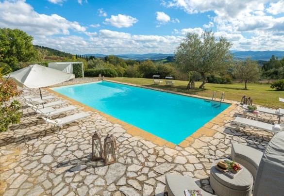 Su una superficie in pietra sono disposti lettini, un divano con poltrone ombreggiati da un gazebo ed un ombrellone – Cinque Cipressi