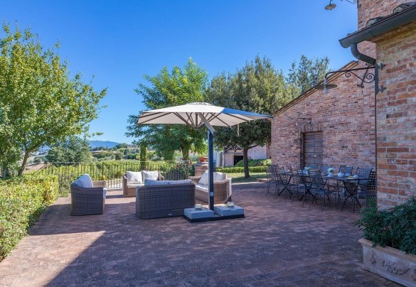 The loggia contains a seating area and dining table covered by large umbrellas, with a stone barbecue – Il Fitto