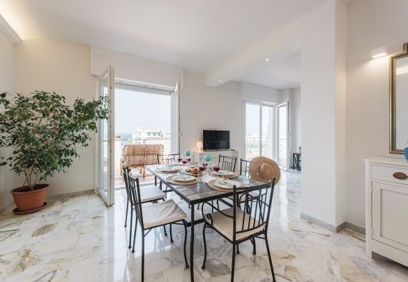 Dall'ingresso di questo attico situato al quinto piano, si accede in un luminoso e largo open space con finestre vista mare che accoglie la cucina e l’area pranzo. Nova Domus