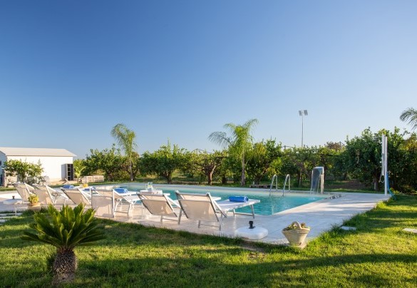 La pavimentazione in gres che profila la piscina accoglie il solarium su cui sono disposti lettini ombreggiati da ombrelloni - Profumo di Zagara