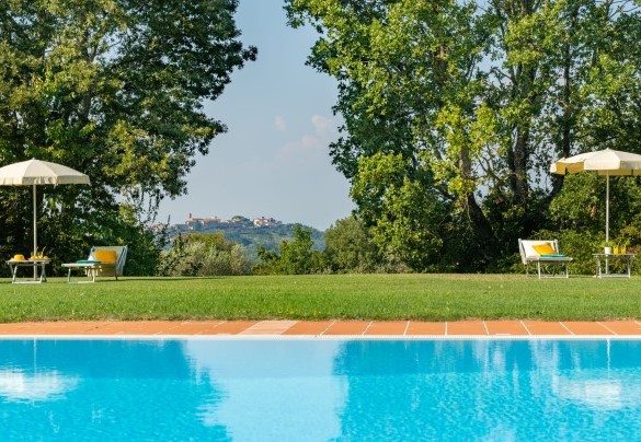 La piscina si trova nel verde di un bellissimo prato circondato dallo svettare del bosco, più in là, oltre il bosco, il borgo di Montefoscoli sullo sfondo – Tenuta di Pratello