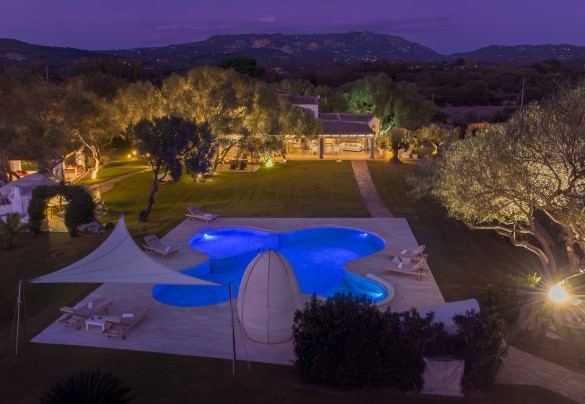 Vista aerea notturna del grande parco privato illuminato, armoniosamente animato da piante autoctone e palme esotiche che ospita la lussuosa piscina interamente rivestita in marmo di Orosei –Villa Typha